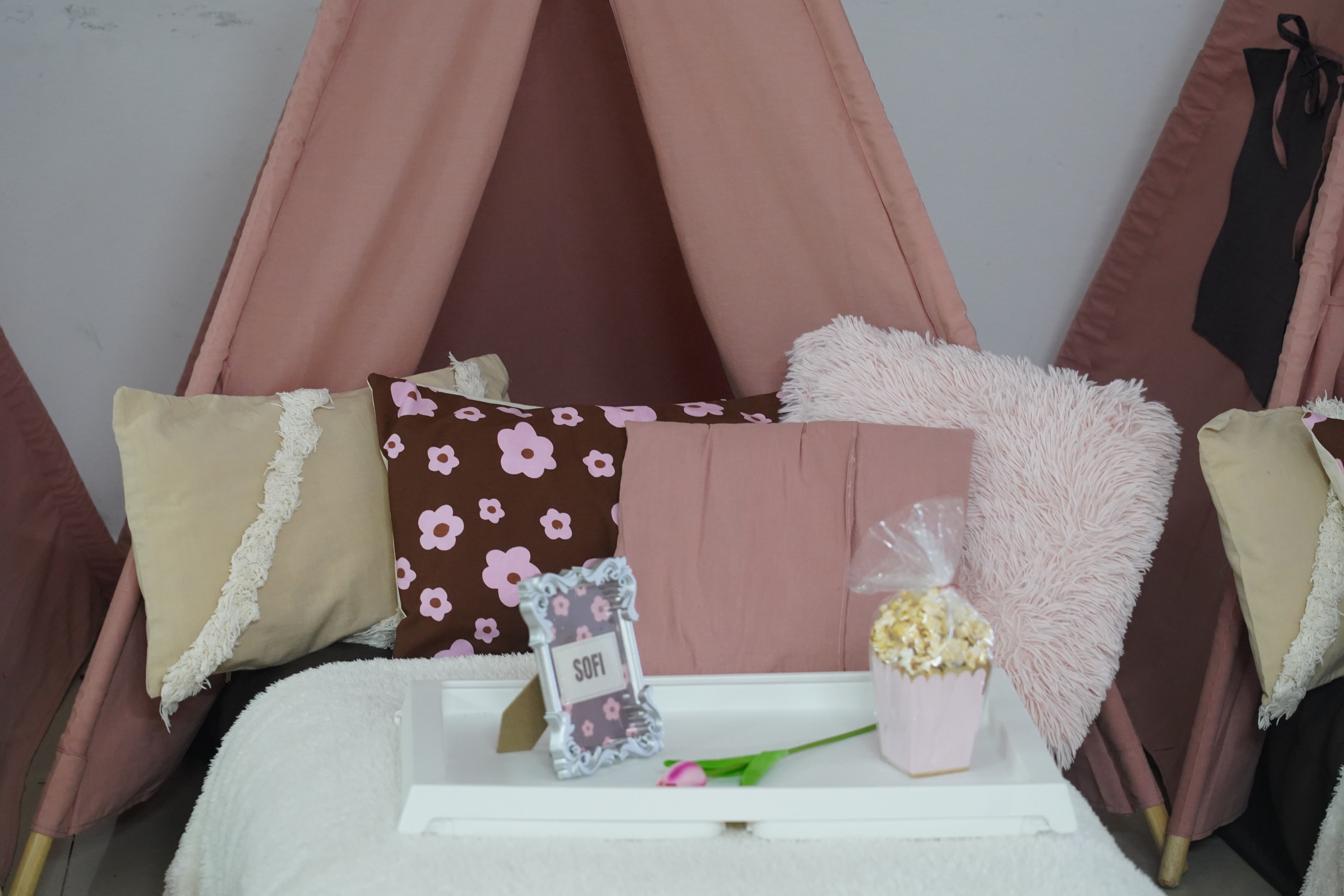 Bandeja blanca con portarretrato, tulipán y pochoclos sobre un colchón, frente a una carpa rosa con almohadones floreados y mullidos.