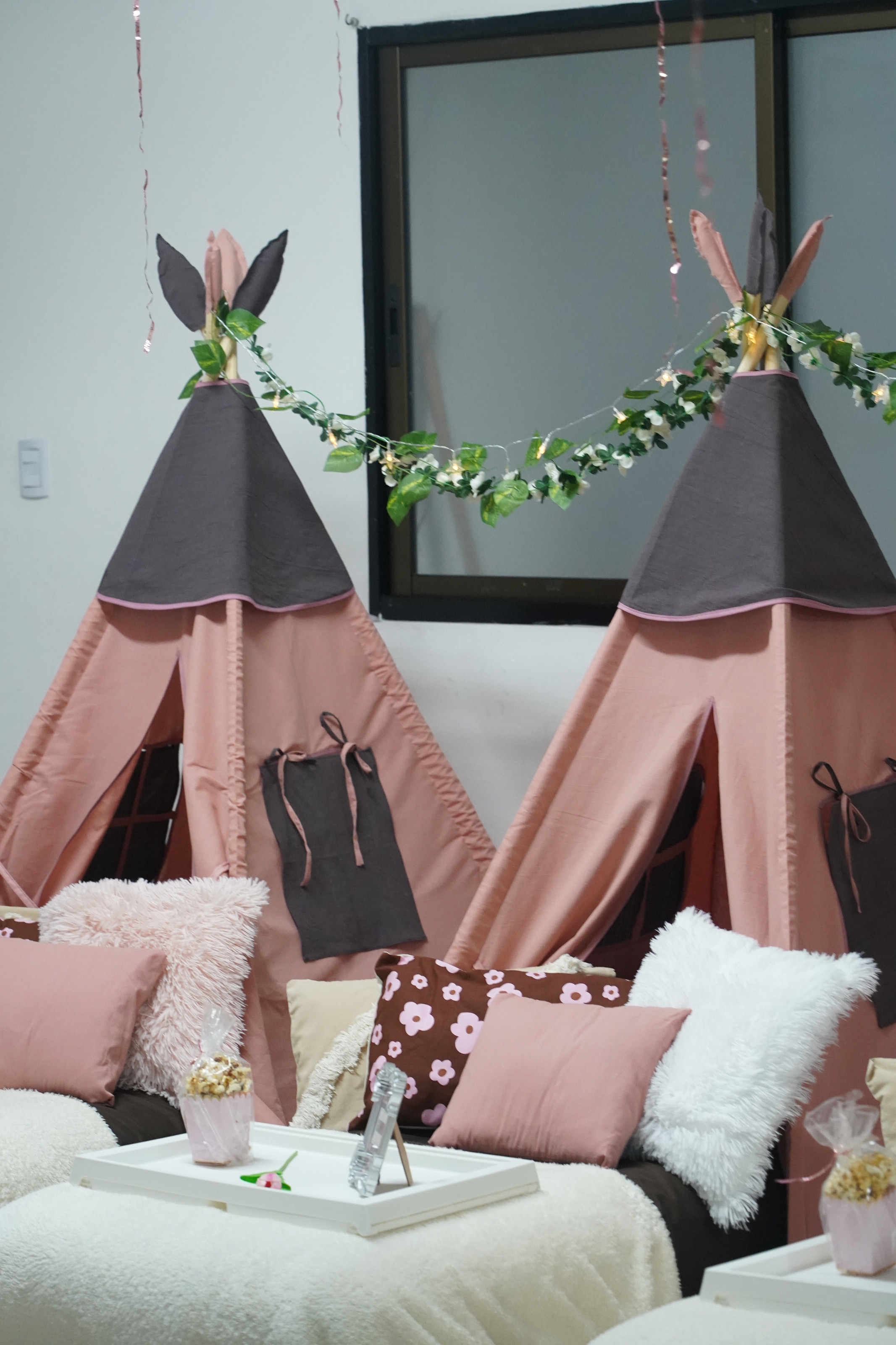 Dos carpas tipi rosa con techo chocolate, unidas por una guirnalda de hojas y luces, con almohadones mullidos y una bandeja con pochoclos.