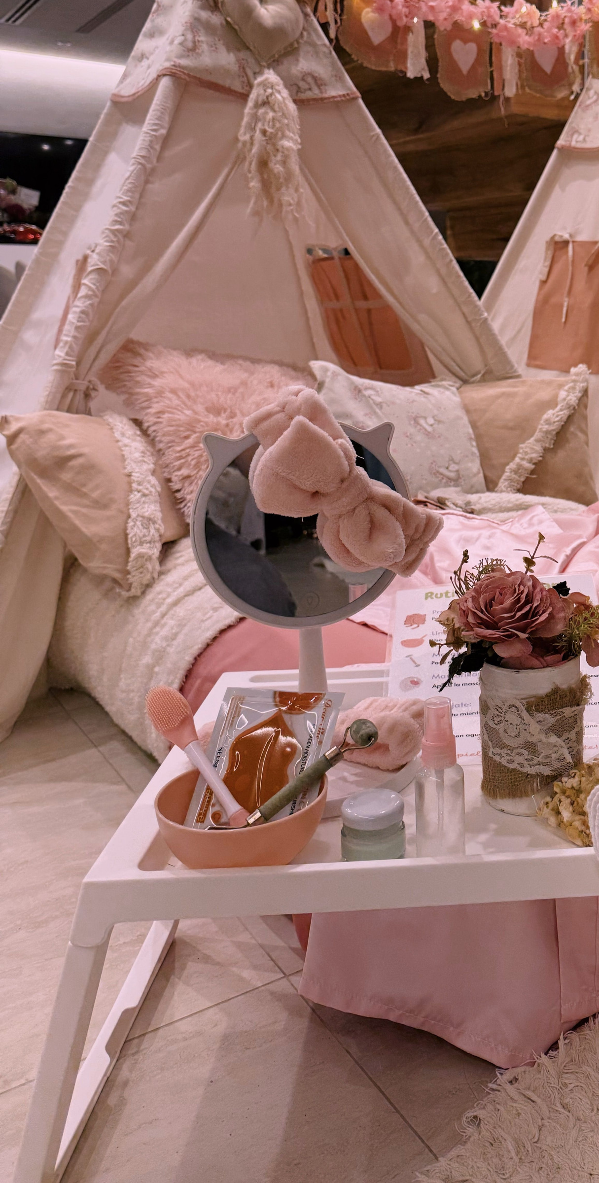 Bandeja del kit de spa con espejo con luces, vincha de moño, bowl, cremas y la rutina de skin care, frente a carpas rosas.